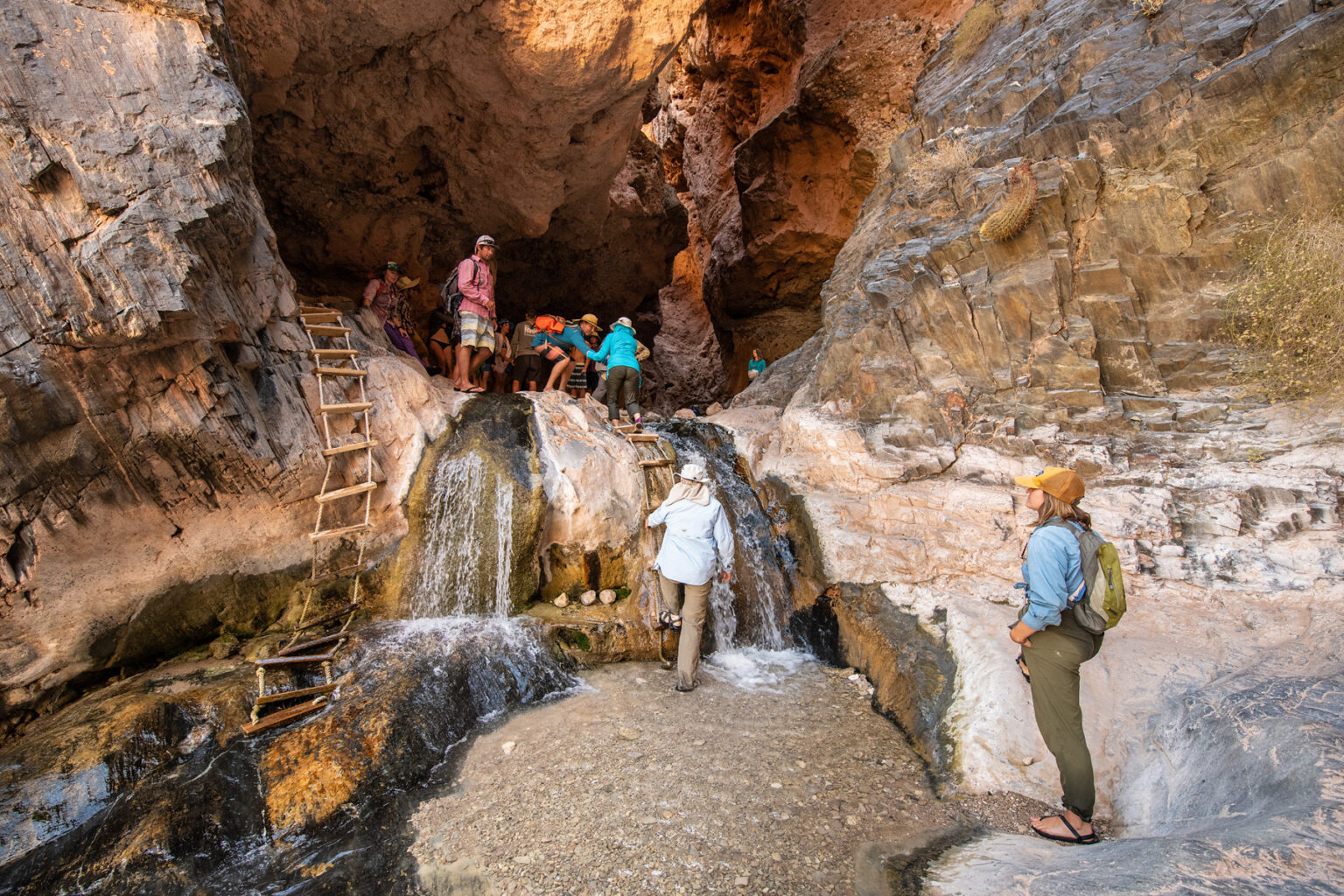 Guests ascend a wood and rope ladder up to a cool cave overlooking a small waterfall in Grand Canyon