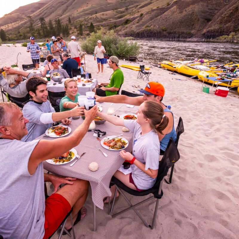 Rafting group sitting at tables, cheersing their craft beers on the bank of the Lower Salmon river.