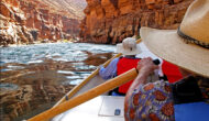 View downstream looking over OARS dory guide's shoulder with guest in PFD and sun hat hanging on in Grand Canyon