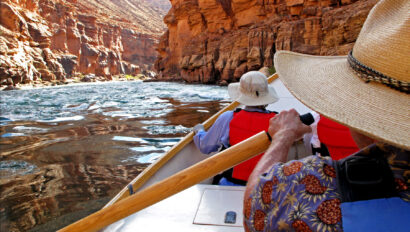 View downstream looking over OARS dory guide's shoulder with guest in PFD and sun hat hanging on in Grand Canyon