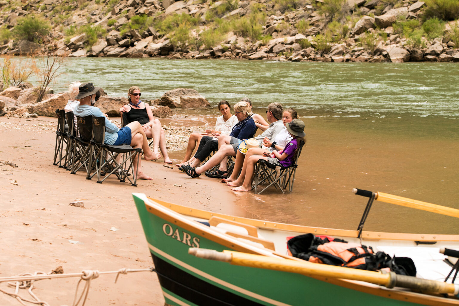 OARS guests chat in a chair circle and the edge of the Colorado River with a dory in the foreground