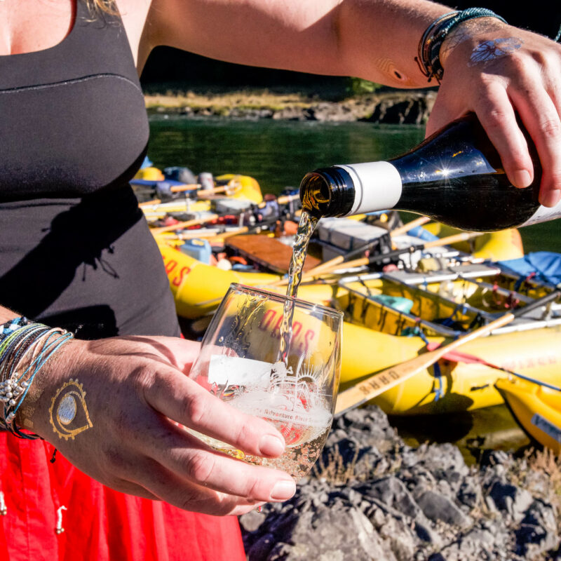 Person pouring wine into a glass on the Rogue River bank.