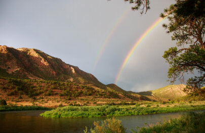 A double rainbow is seen above a riverscape with mountains studded with lots of green vegetation.