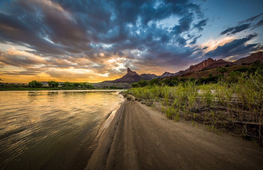 Riverside view of a glorious cloudy sunset over mountains and the river lined with green vegetation.