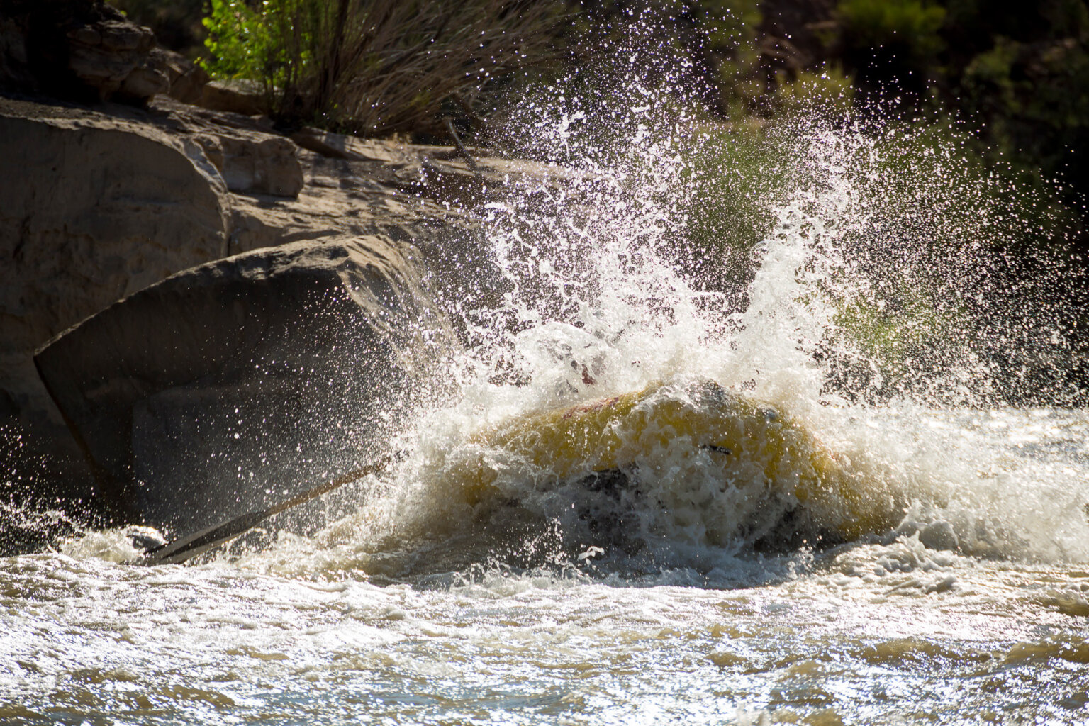 A yellow raft crashes through a rapid and water sprays up all around it obscuring the people sitting in the boat.