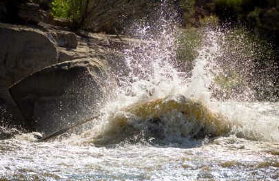 A yellow raft crashes through a rapid and water sprays up all around it obscuring the people sitting in the boat.