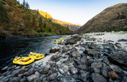 A wide view of camp set up along the shoreline of the Lower Salmon River and several yellow rafts are tied up with tents and the chair circle in the back.