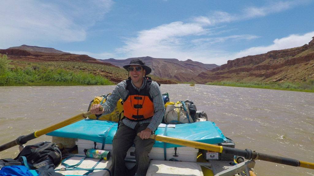A man rows a raft on the San Juan River, surrounded by red cliffs