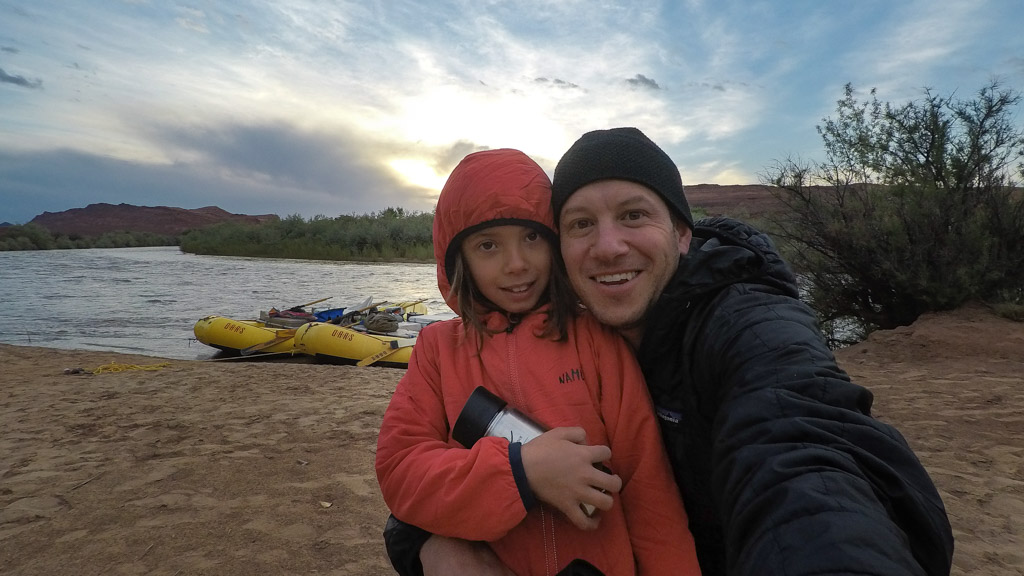 A father and daughter wearing coats pose for a selfie on the shore of the San Juan River