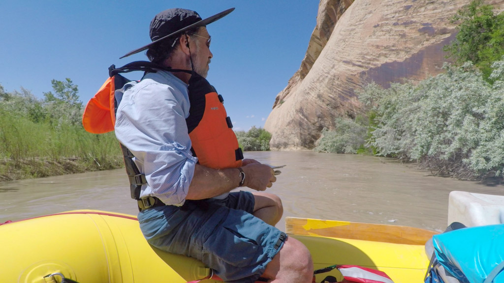 An older man in a lifejacket balances on the edge of a raft in a calm stretch of river
