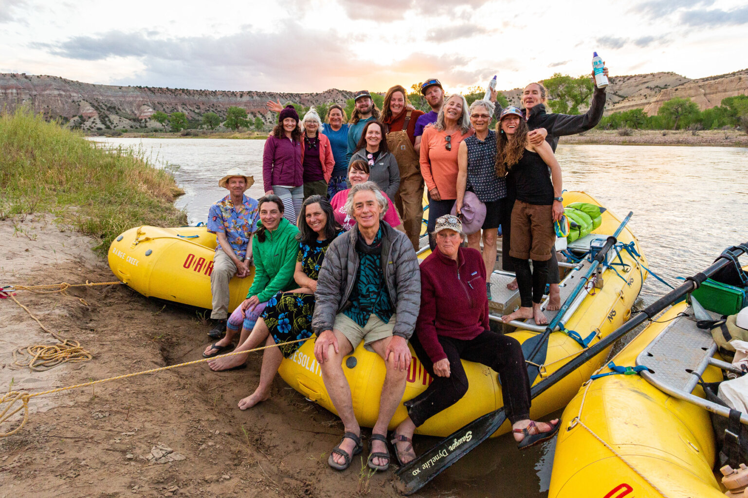 A group of smiling guests and guides sit and stand on yellow rafts tied up along the shoreline for a group photo on a Yampa River trip.