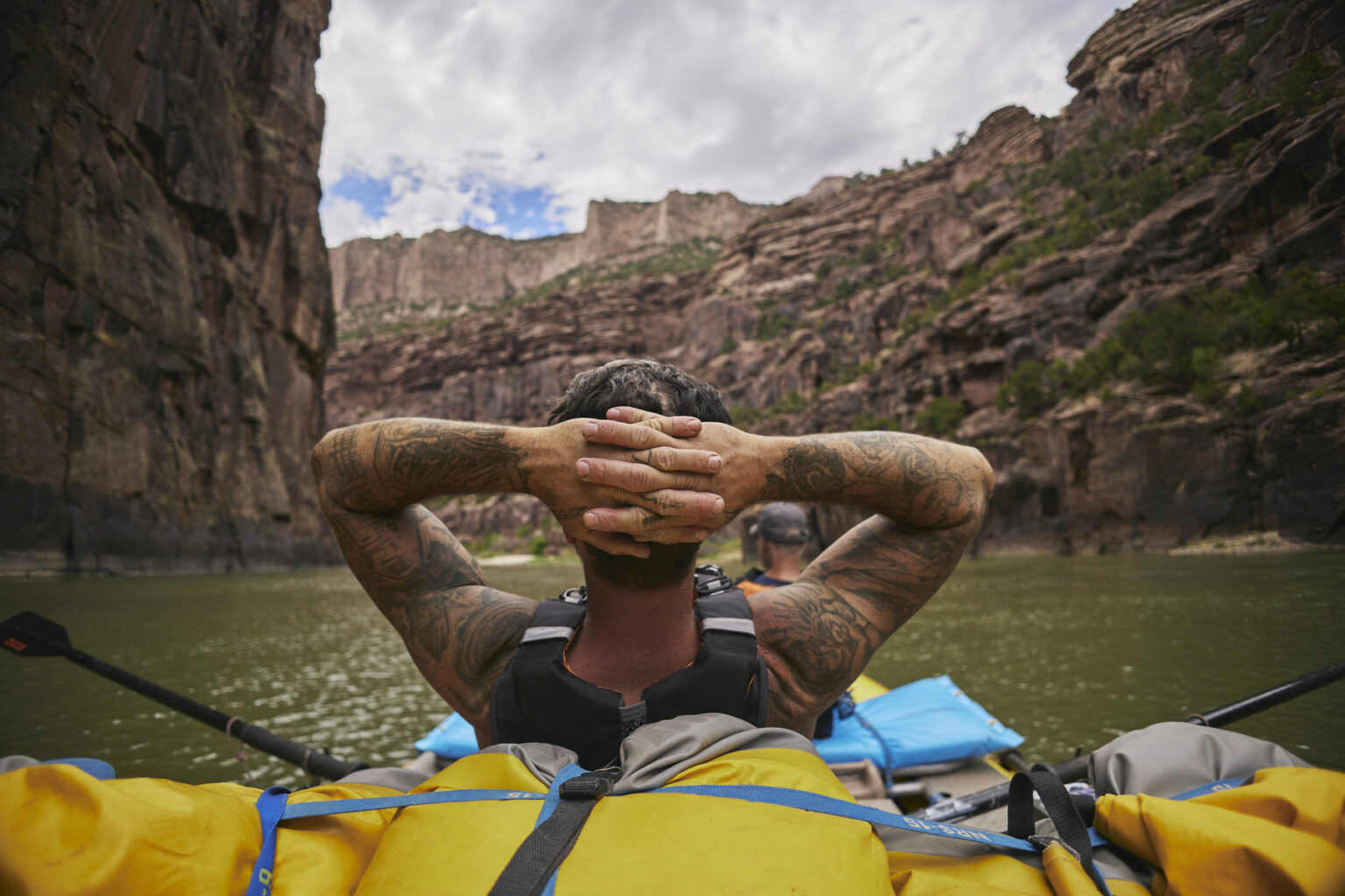 A tattooed guide leans back cradling the back of his head in his hands and takes in the beauty of the Gates of Lodore on the Green River in Utah.