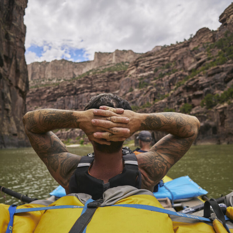 A tattooed guide leans back cradling the back of his head in his hands and takes in the beauty of the Gates of Lodore on the Green River in Utah.