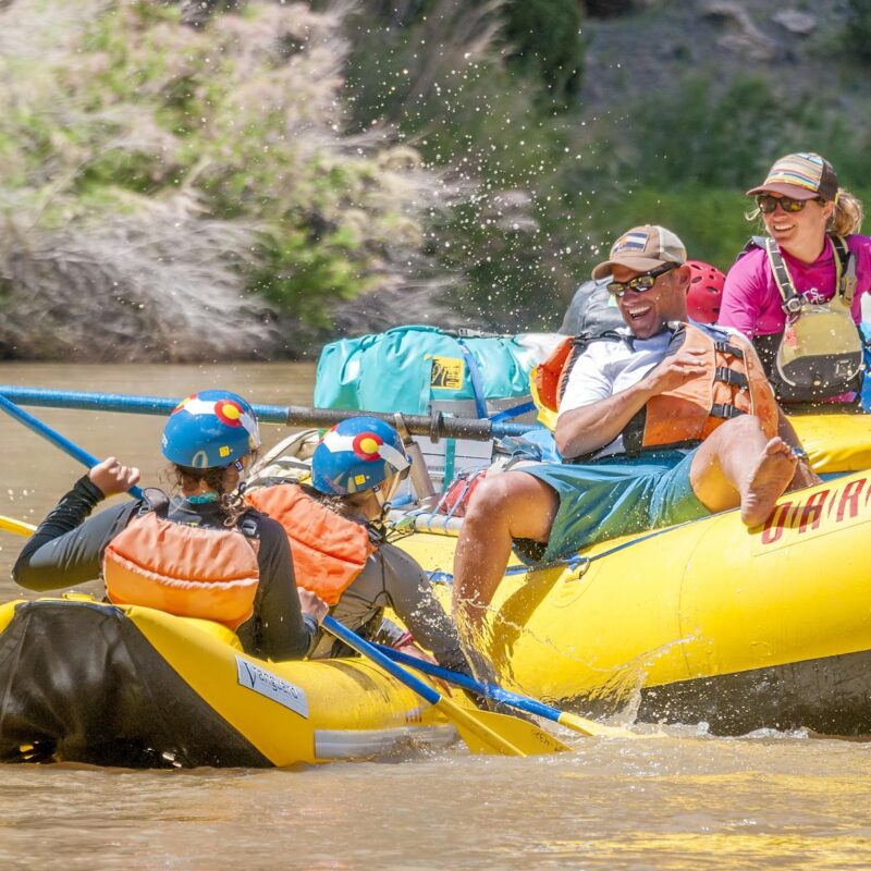 Three guests engage in a fun water fight on the Yampa River while a guide watches and smiles.