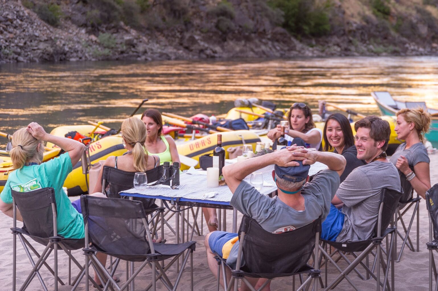 A group of adults sit around a linen draped table on a sandy beach drinking wine and socializing on an OARS Wine on the Salmon River trip.
