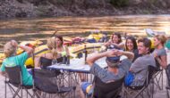 A group of adults sit around a linen draped table on a sandy beach drinking wine and socializing on an OARS Wine on the Salmon River trip.