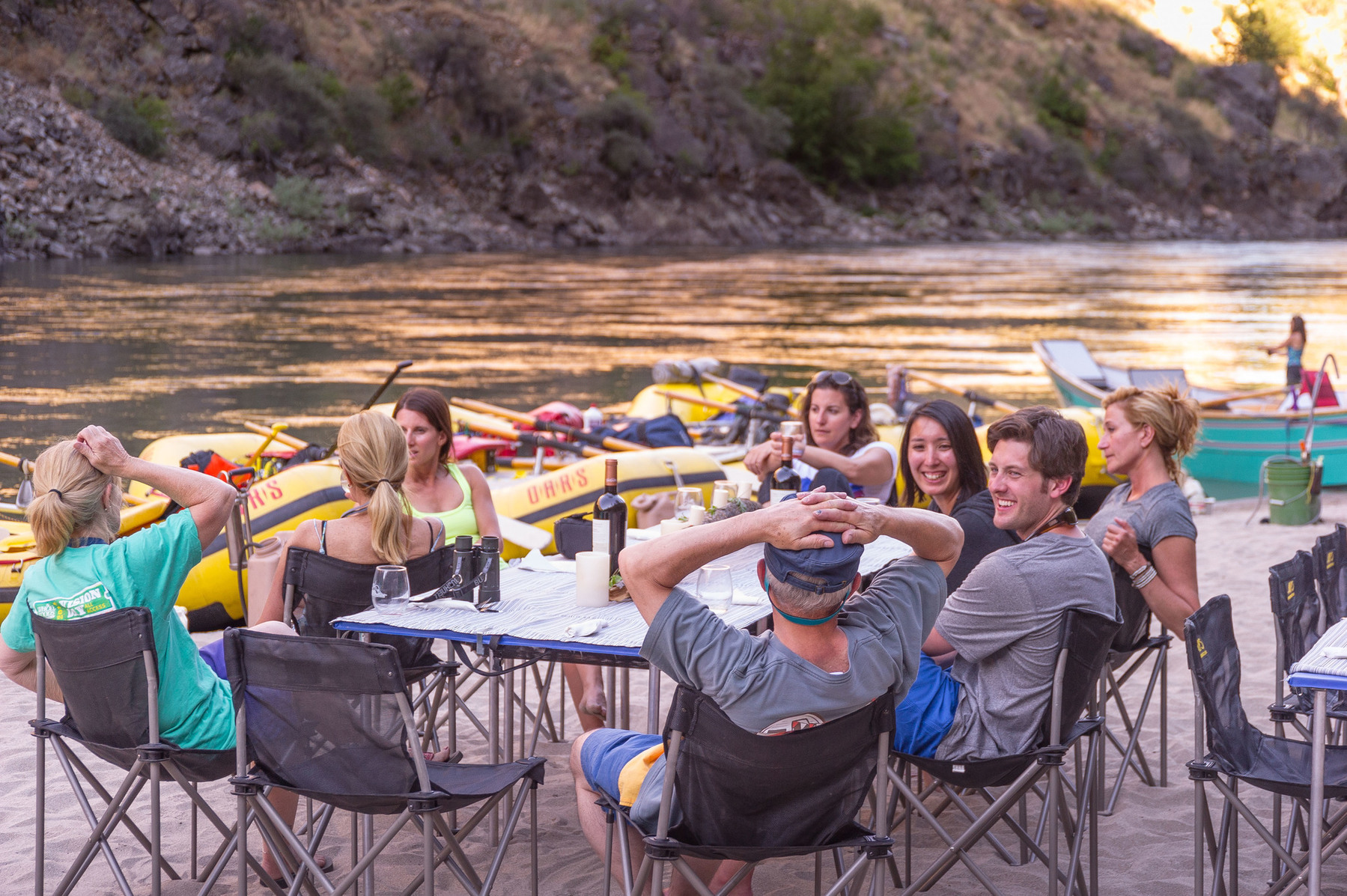 A group of adults sit around a linen draped table on a sandy beach drinking wine and socializing on an OARS Wine on the Salmon River trip.