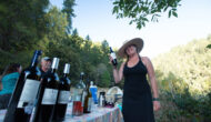 A smiling woman holds up a bottle of wine as guests look on enthusiastically on the other side of a table lined withvarious wines on a Wine on the Rogue River trip.