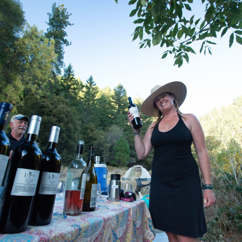 A smiling woman holds up a bottle of wine as guests look on enthusiastically on the other side of a table lined withvarious wines on a Wine on the Rogue River trip.