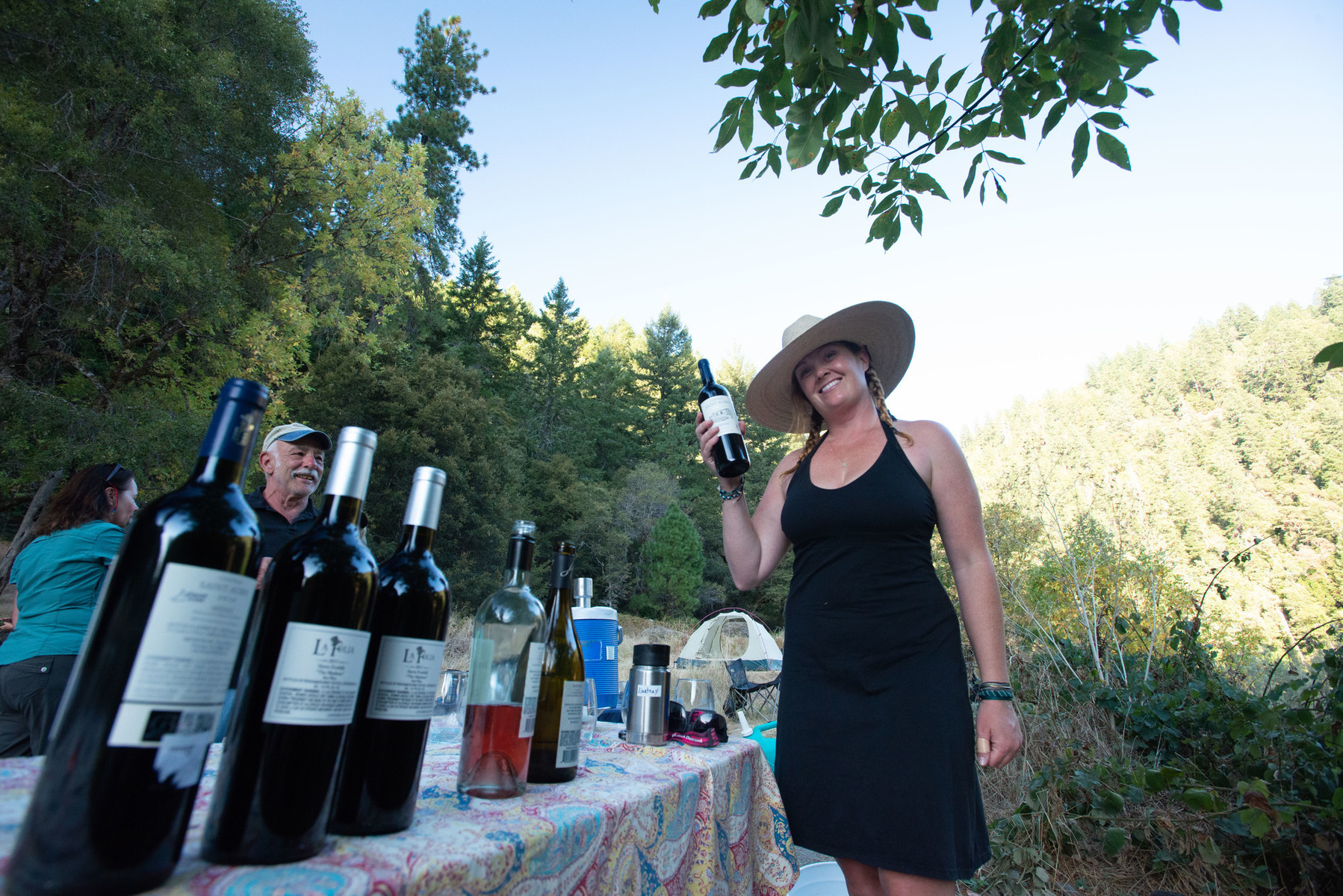 A smiling woman holds up a bottle of wine as guests look on enthusiastically on the other side of a table lined withvarious wines on a Wine on the Rogue River trip.