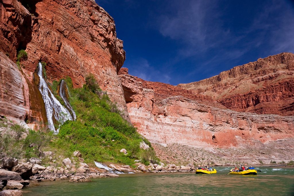 Two yellow rafts float down the Colorado River with Vasey's Paradise, a natural spring in Grand Canyon, gushing water off the cliffside