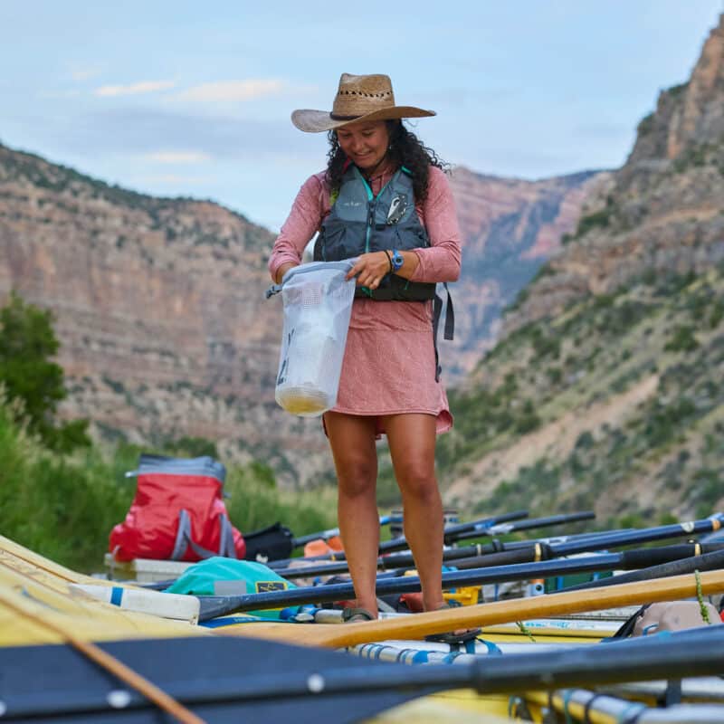 woman in a hat reaching into a bag while standing on a raft on a river.