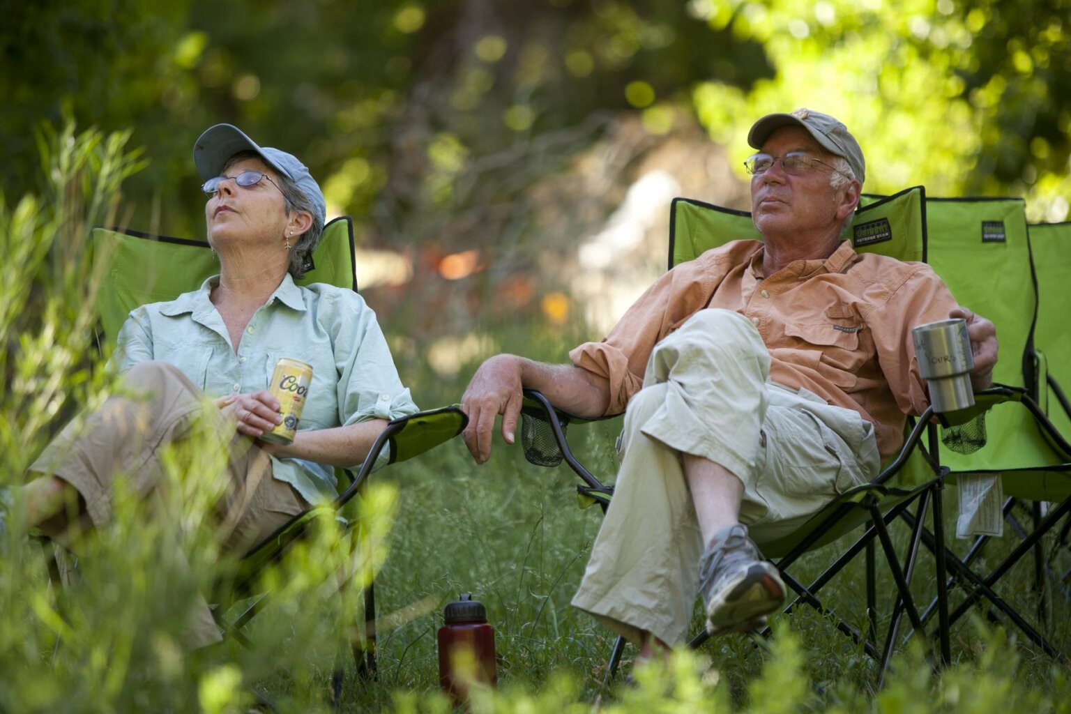 Two guests lean back in camp chairs with beverages and look up at the scenery at their verdant campsite along the Yampa River in Utah.