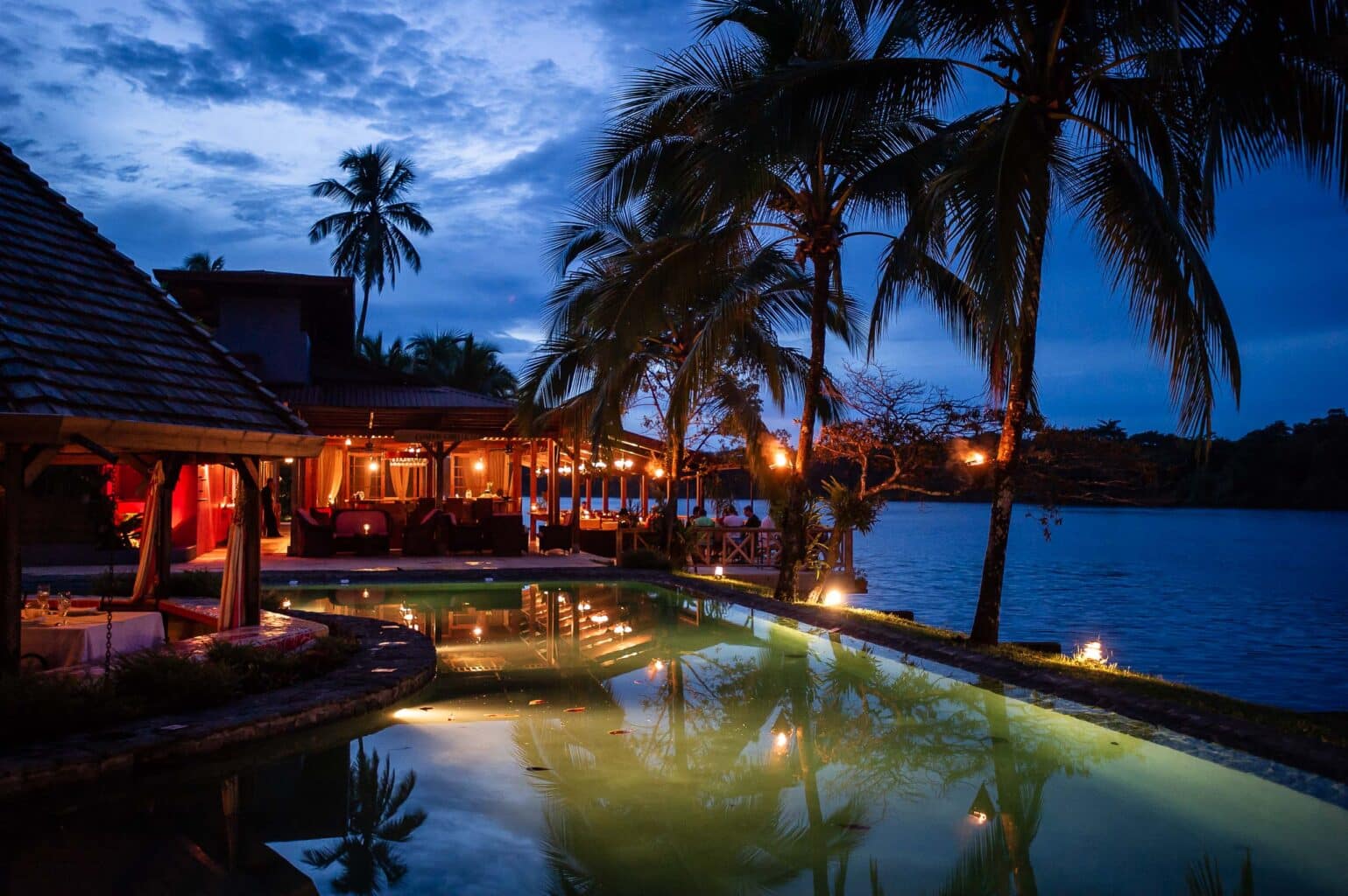 lodge with pool under palm trees next to a lake at dusk.