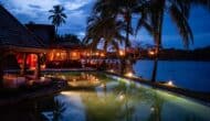 lodge with pool under palm trees next to a lake at dusk.