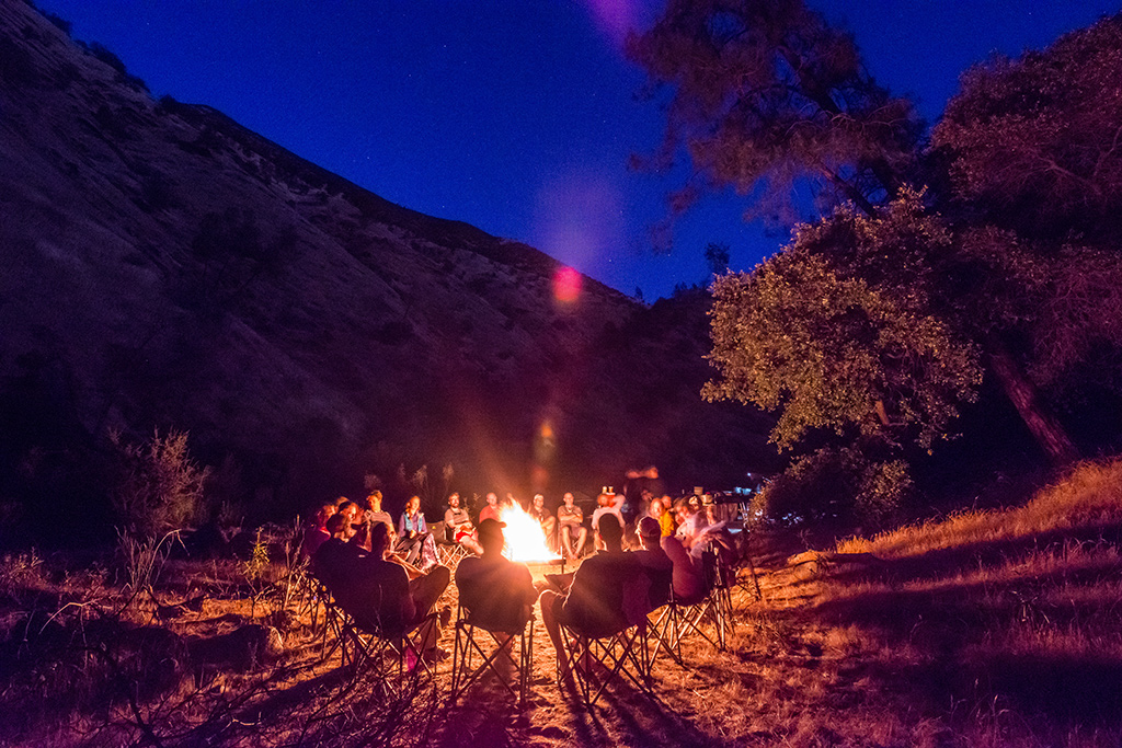 A rafting bachelor party group sits around a campfire in the chair circle at night on the Tuolumne River in California