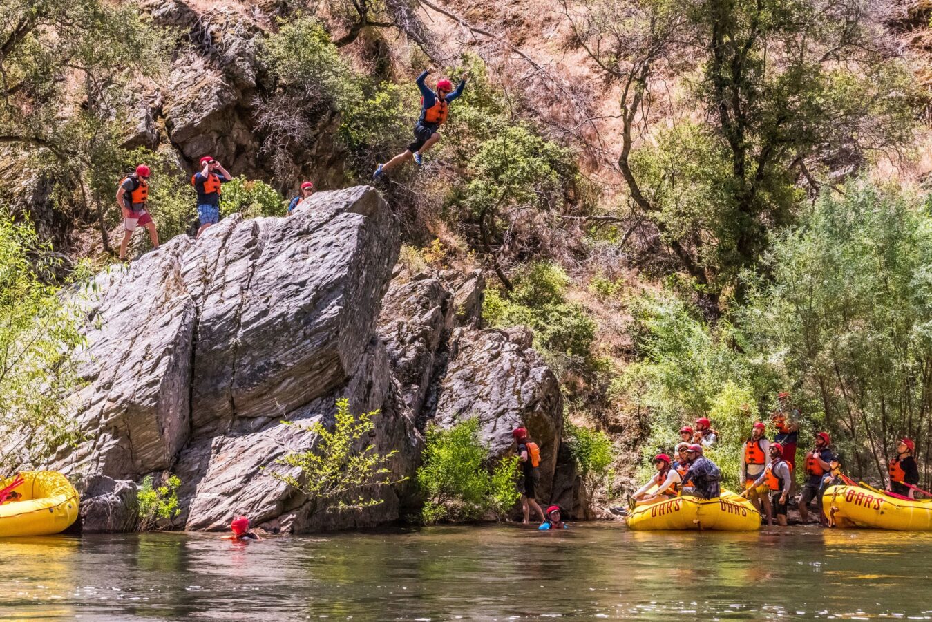 A person mid-air jumping off a rock into the Tuolumne River as a group of guests watches