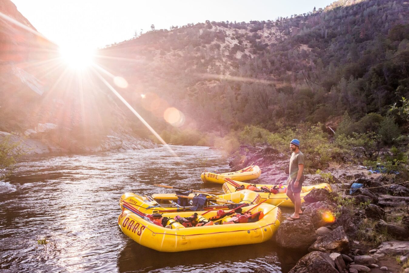 Four yellow OARS rafts tied up at camp while a man stands on a rock with a sunburst over the mountains on the Tuolumne River