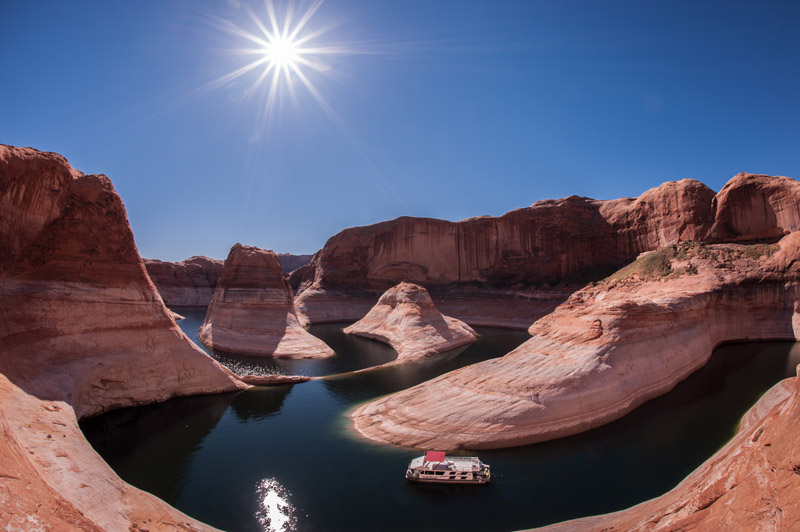 A houseboat sits in the green water and high orange cliffs of Lake Powell, Reflection Canyon