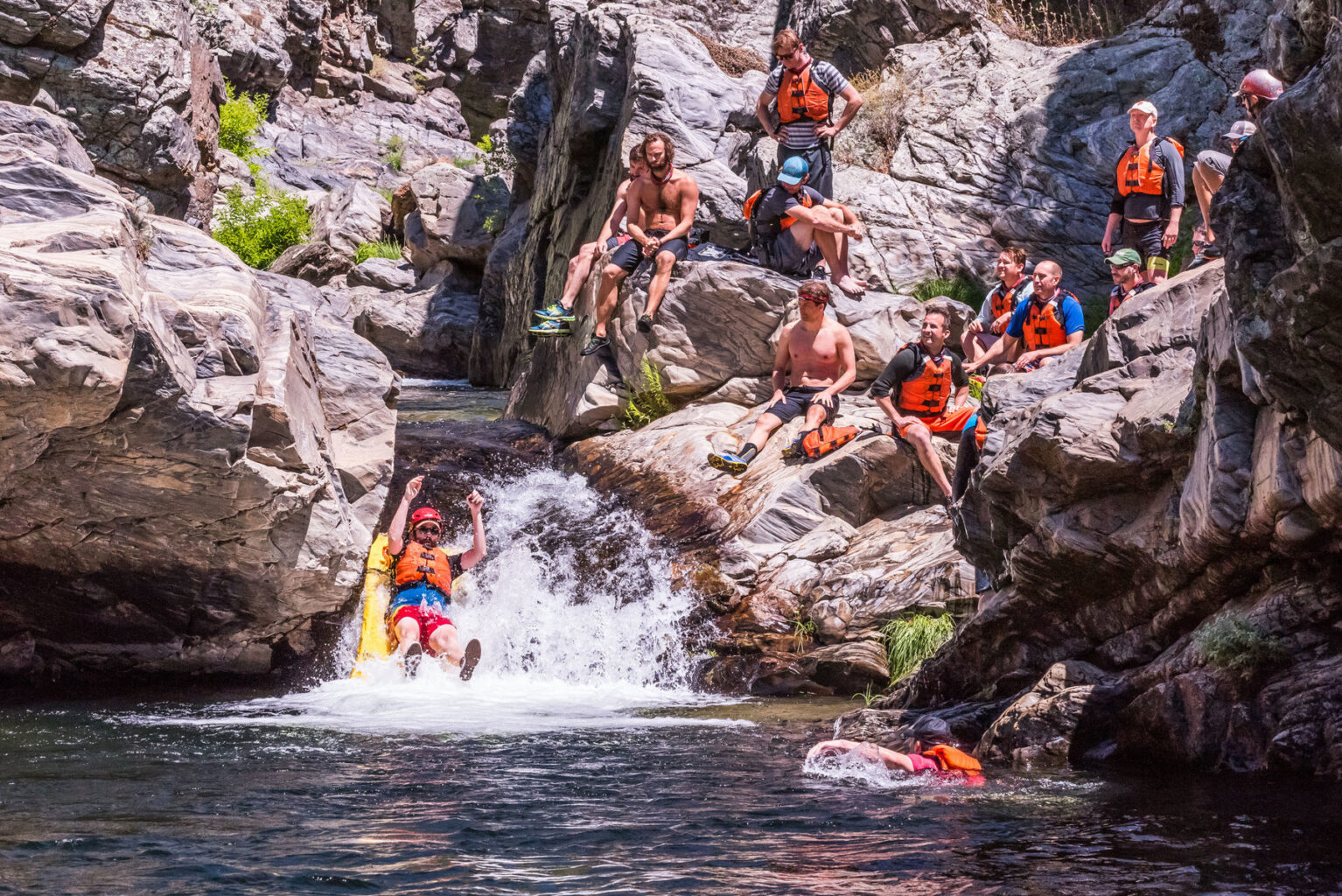 A man going down a natural waterslide while a group of men sit on the rocks and watch.