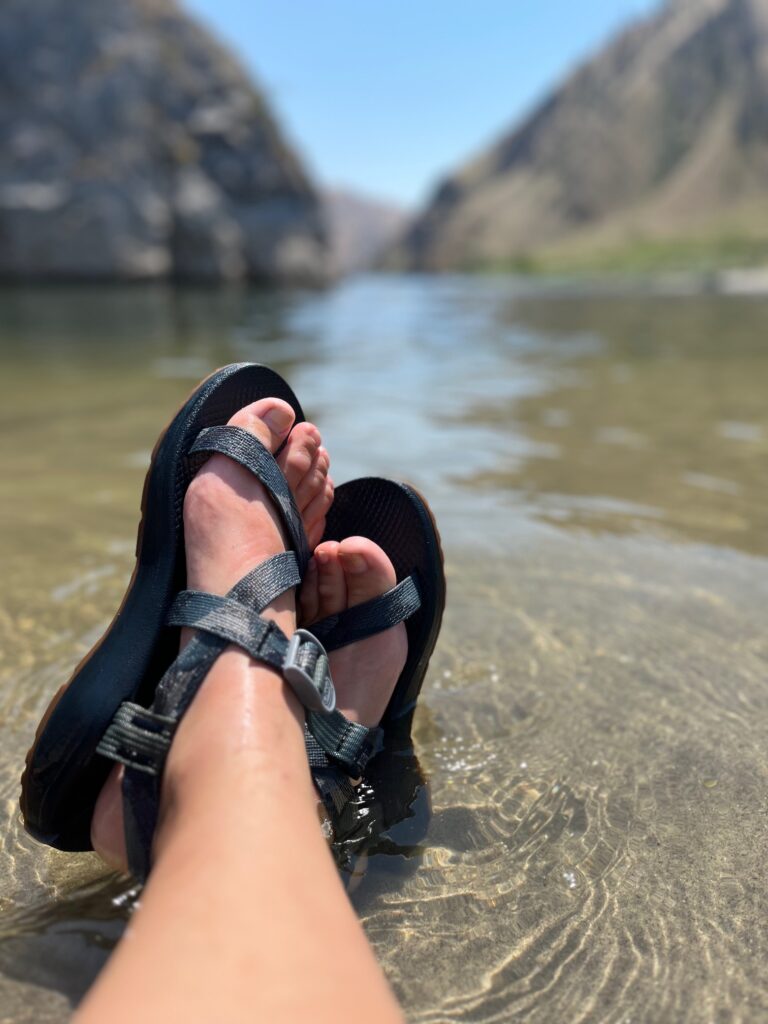 Feet with river sandals wading in a river with scenic canyon in the background