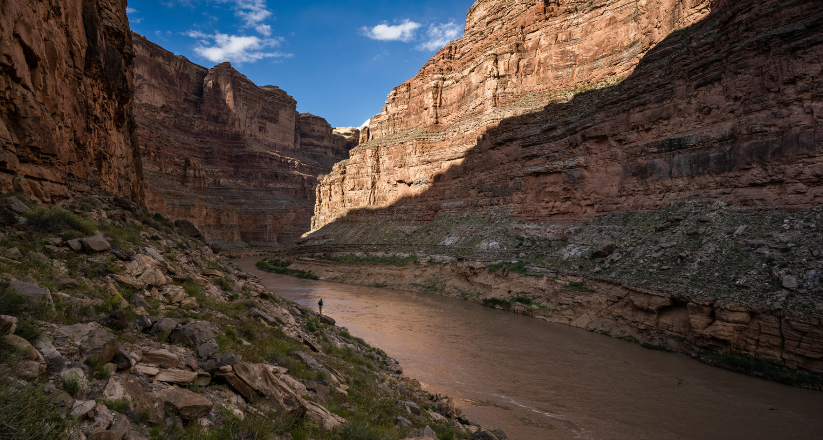 A lone person stands along the the Colorado River with the walls of Cataract Canyon towering above them