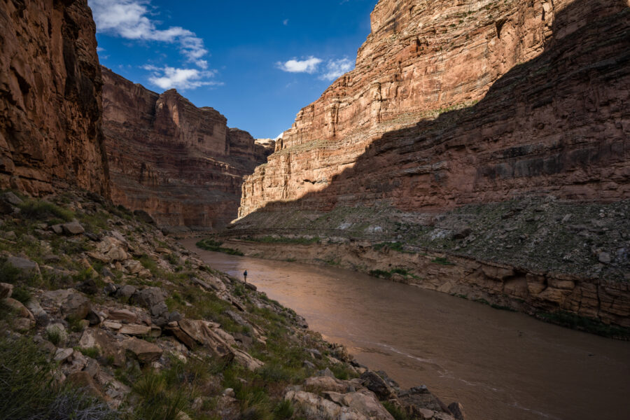A lone person stands along the the Colorado River with the walls of Cataract Canyon towering above them