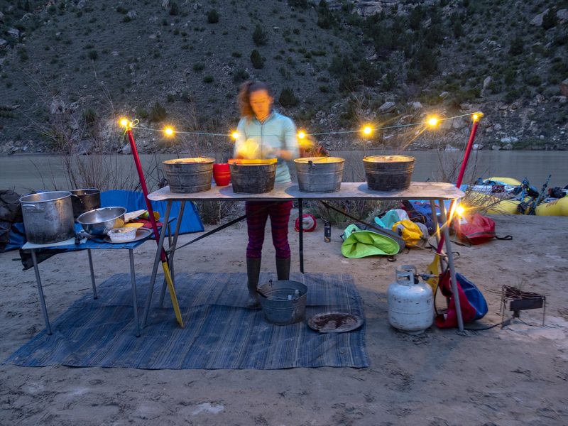 An OARS guide washes dishes under string lights on a rafting trip in Utah