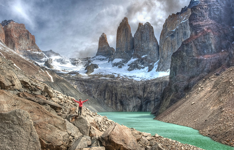 A hiker stands on a boulder in the distance with arms out to their side as they pose in front of rising mountains along a turquoise green river in Patagonia.
