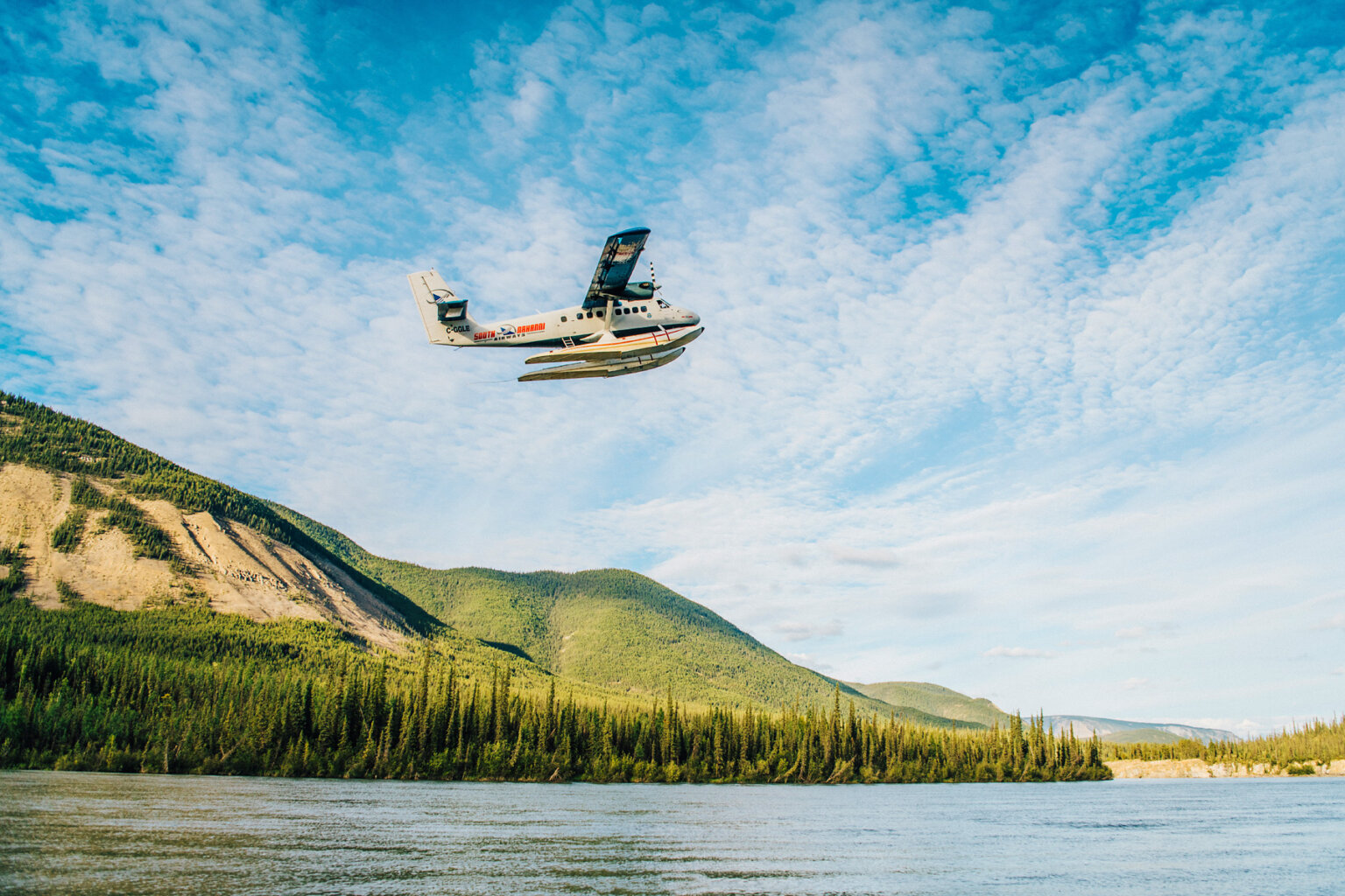 A water plane flies over the river as it takes off surounded by lush, green forested hills.