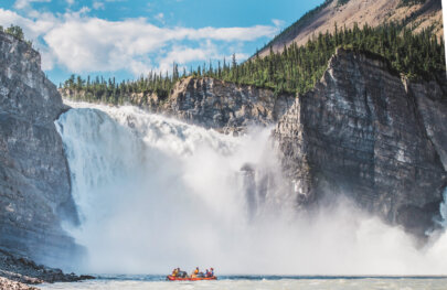 A raft full of people sits on the water in front of a massive, wide waterfall on the Nahanni River in Alaska.