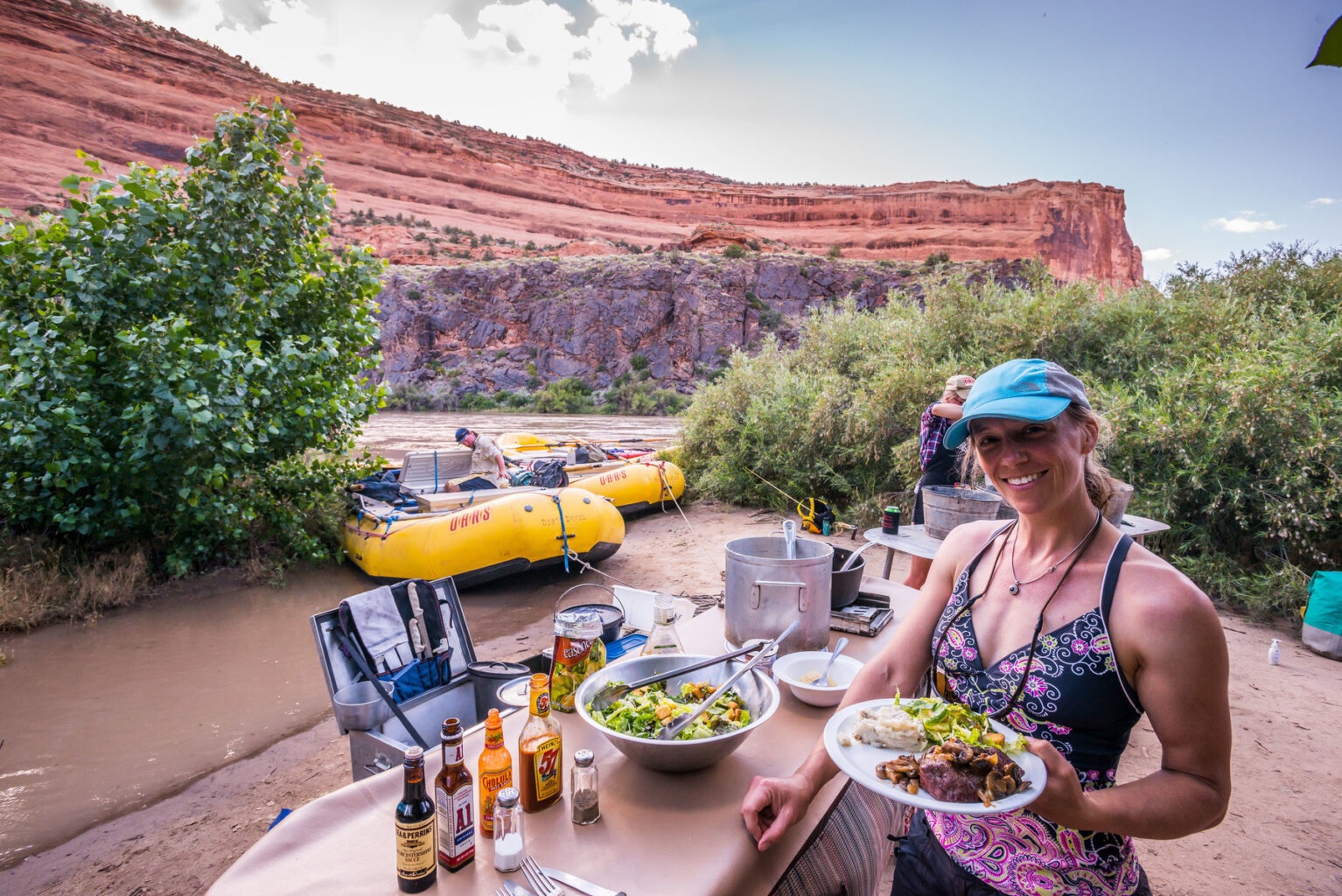 A guest smiles at the camera and holds up a plate of food next to the buffet line for dinner at camp along the Colorado River in Westwater Canyon.