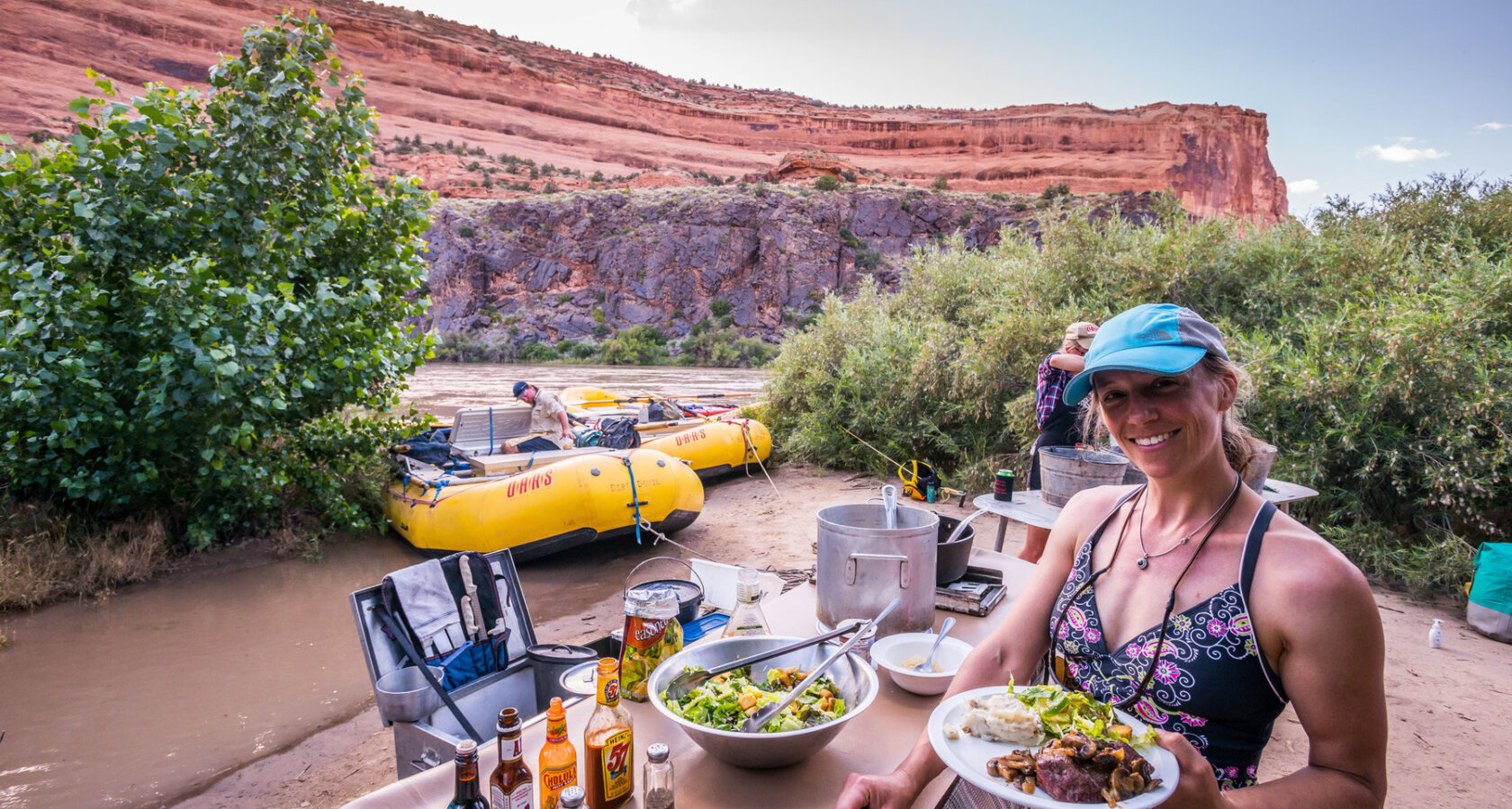 A guest smiles at the camera and holds up a plate of food next to the buffet line for dinner at camp along the Colorado River in Westwater Canyon.