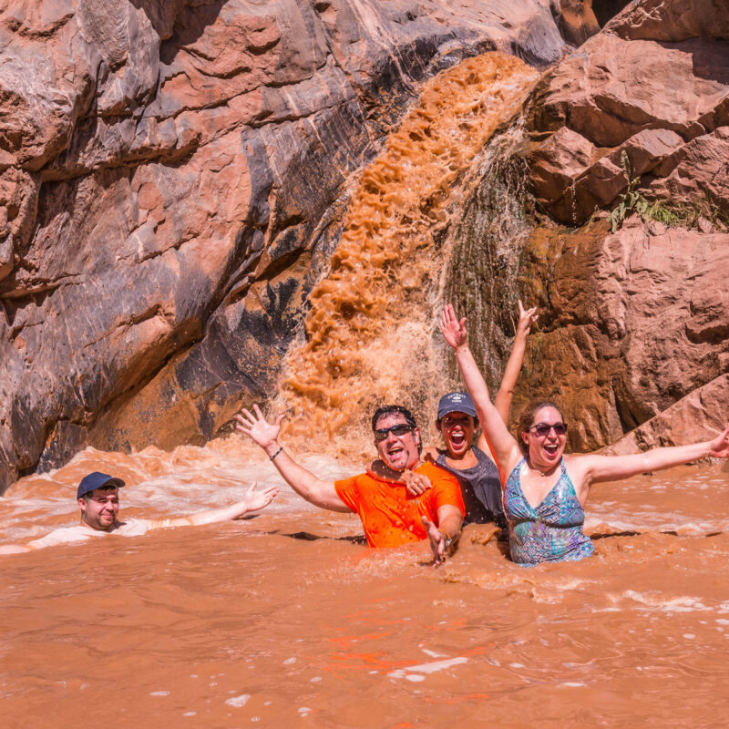 Four adults playing in the Colorado River in front of a waterfall in Westwater Canyon as they smile at the camera with their hands in the air