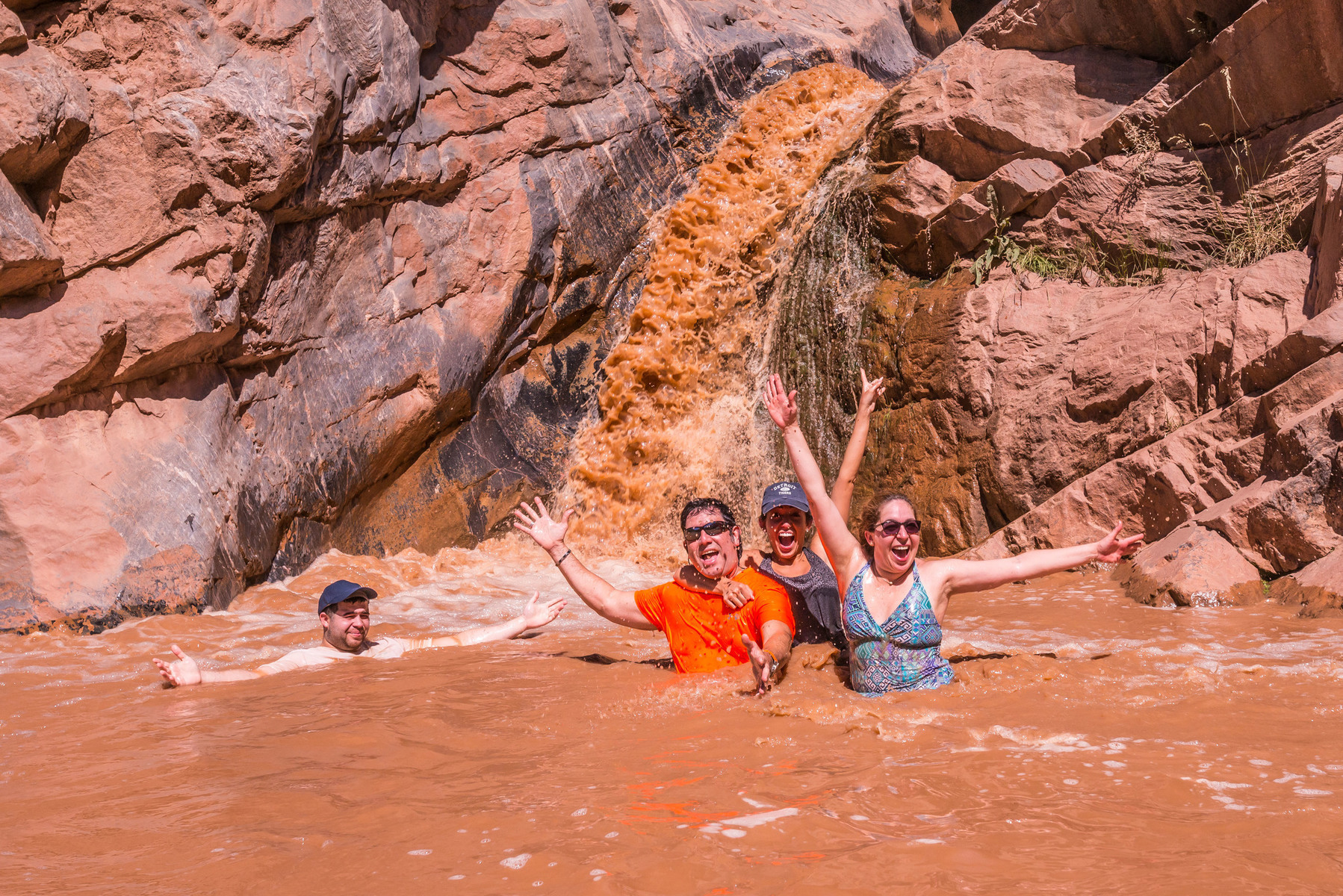 Four adults playing in the Colorado River in front of a waterfall in Westwater Canyon as they smile at the camera with their hands in the air