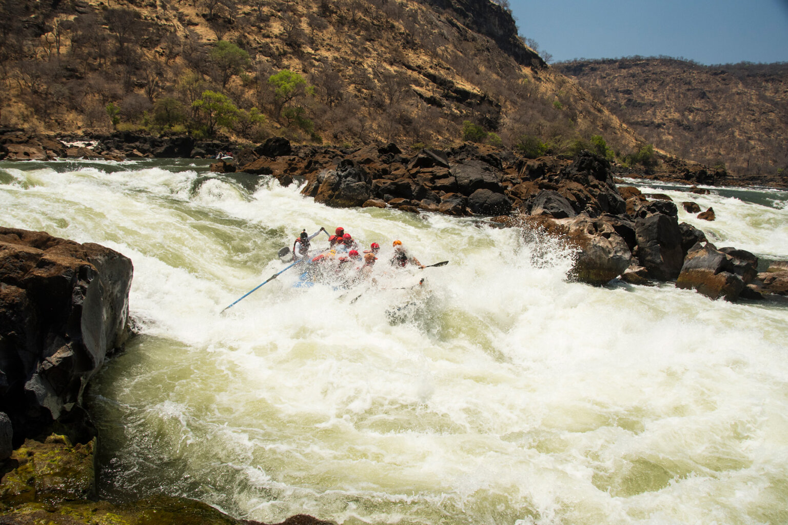 A raft full of people as it is nearly fully submerged by a wave of whitewater as a guide steers on oars through a huge rapid on the Zambezi River in Africa.