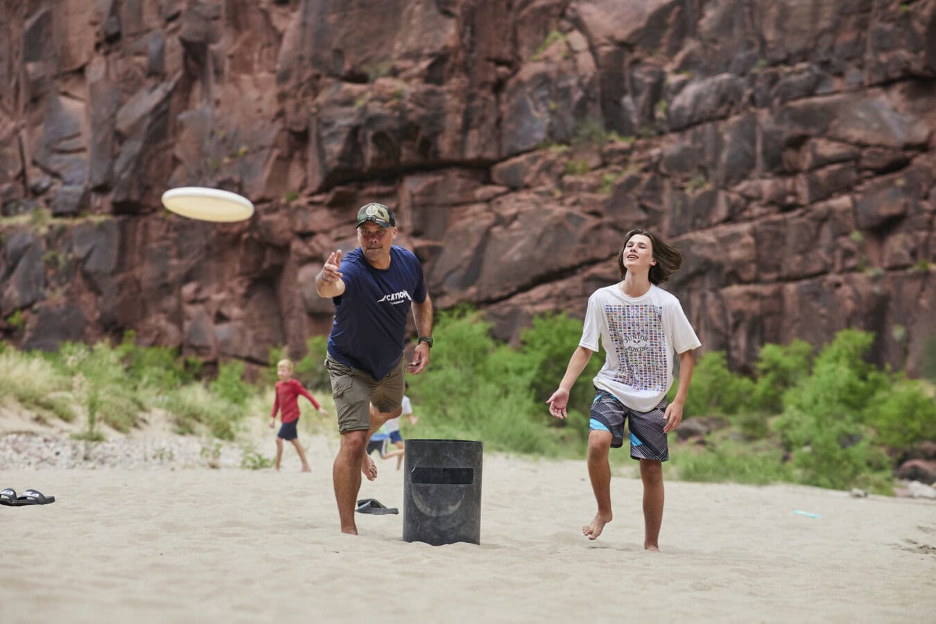 A man and an older child play a frisbee game on a sandy beach souorronded by rocky cliffs and greenery