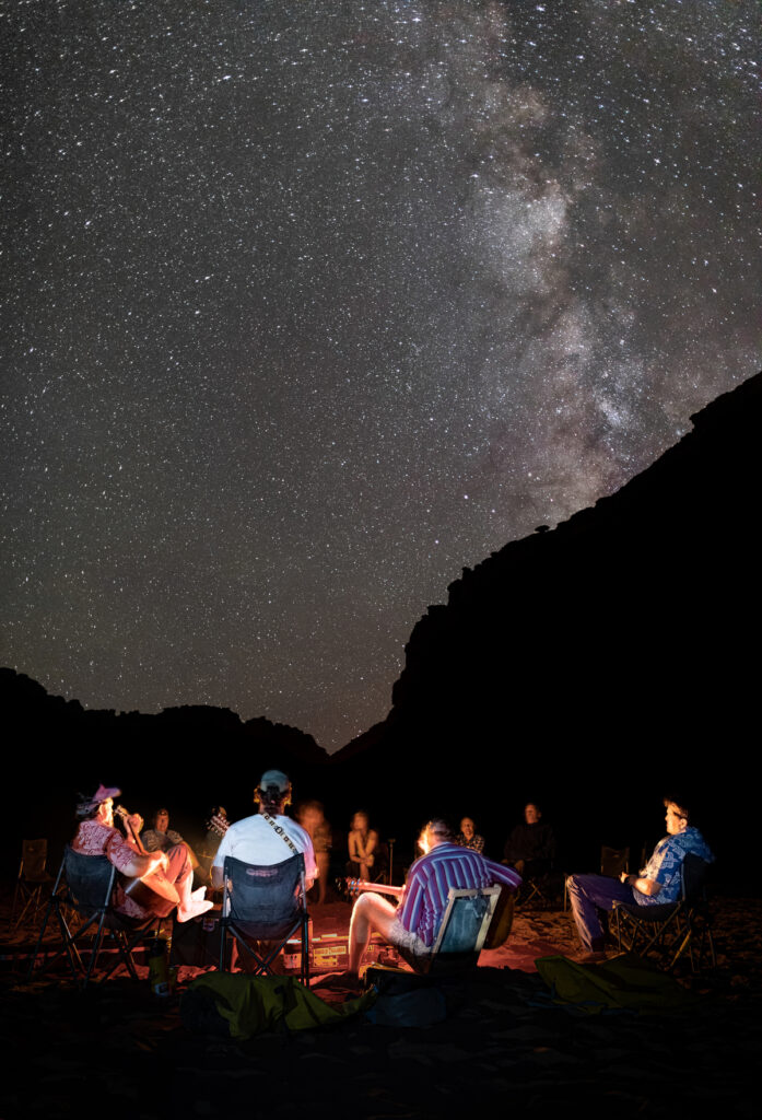 Three musicians play their instruments to a group huddled around a campfire under a starry night sky in Canyonlands National Park