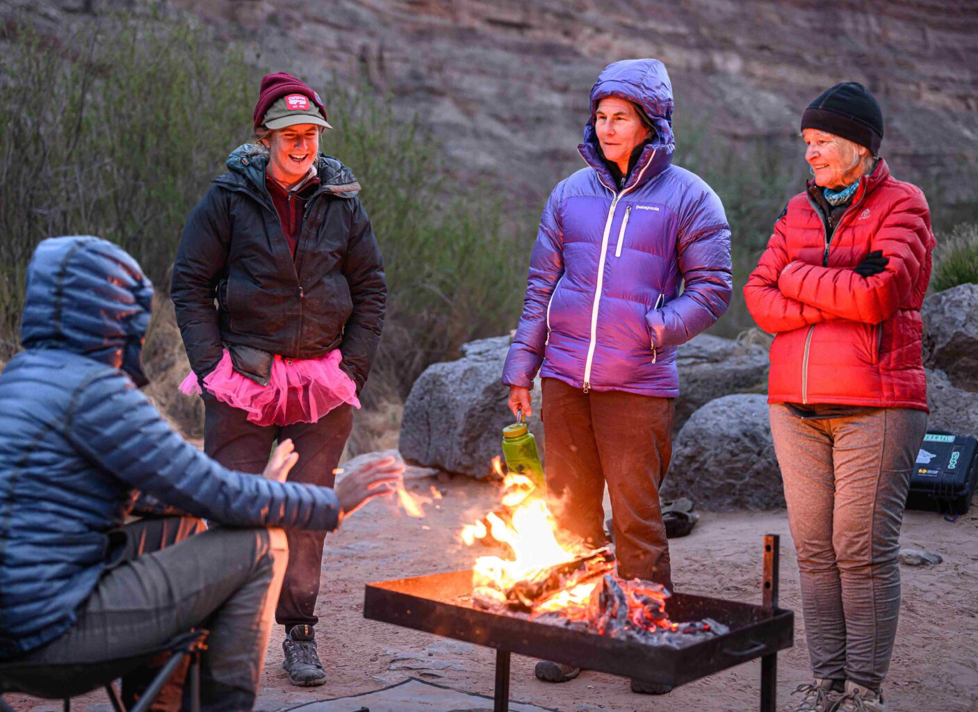 A group of OARS guests and guides stand around a camp fire on a cold rafting trip