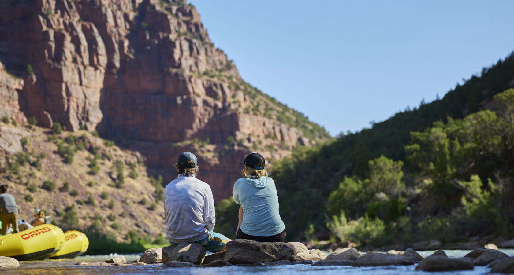 A couple sitting on a rock near a river gazing off at a towering red rock canyon wall